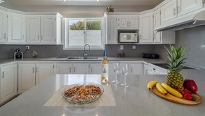 Une cuisine moderne avec des meubles blancs, un grand îlot central sur lequel on trouve un bol de fruits secs, une bouteille de vin et une assiette de fruits frais.