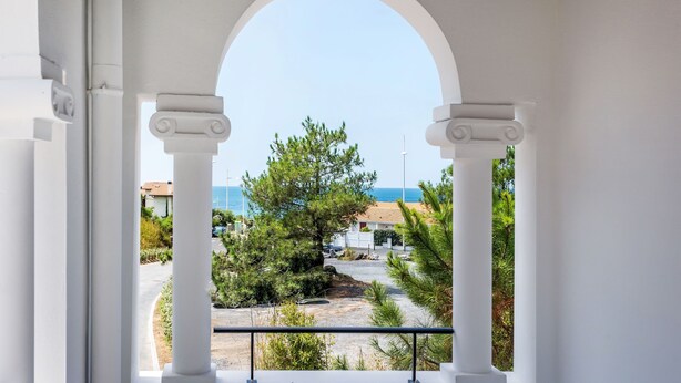 Une vue à travers une porte en arc surmontée de colonnes blanches, donnant sur une zone côtière avec des arbres et un bâtiment.