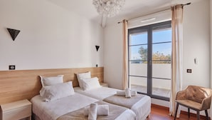Une chambre d’hôtel avec deux lits, une tête de lit en bois, un lustre, une vue par une grande fenêtre et un fauteuil.