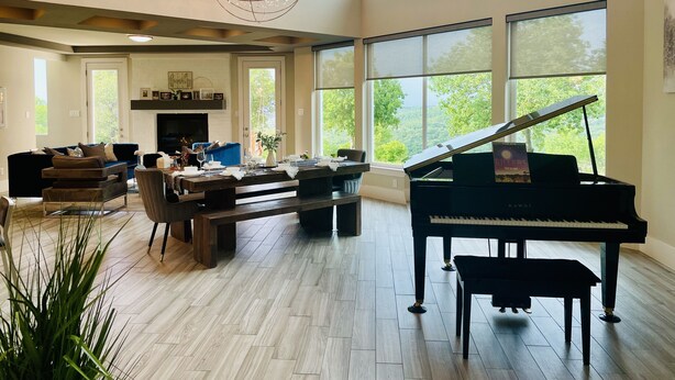 A spacious living room with a grand piano, a chandelier, and large windows offering a view of the outdoors. - Eagles' View Retreat! Family-sized home in Texas countryside with stunning views (Gordon)