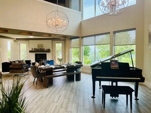 A spacious living room with a grand piano, a chandelier, and large windows offering a view of the outdoors. - Eagles' View Retreat! Family-sized home in Texas countryside with stunning views (Gordon)