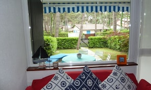 Une pièce donnant sur un jardin, une fenêtre ornée d’une lambrequin à rayures, une petite table avec un bol, et un coussin aux motifs rouges et blancs.