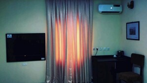 A room with a flat-screen TV mounted on the wall, a ceiling air conditioner, a desk with a chair, and a curtain with a warm light effect. - The 401 Hotel and Suites Awka (Awka)