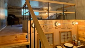 Interior de una cabaña de madera con una cama litera, una escalera y un pequeño rincón de estar.