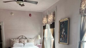 Un dormitorio con una cama, un sofá gris, un ventilador de techo, una ventana con cortinas y un cuadro en la pared.