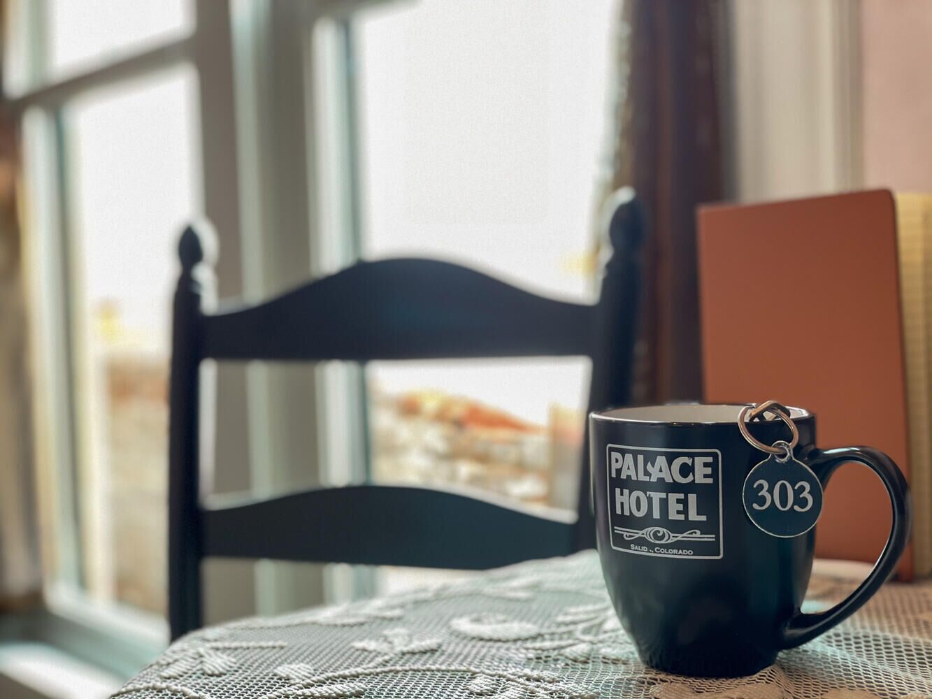A black mug with "Palace Hotel" and "303" on it, placed on a table with a lace tablecloth.