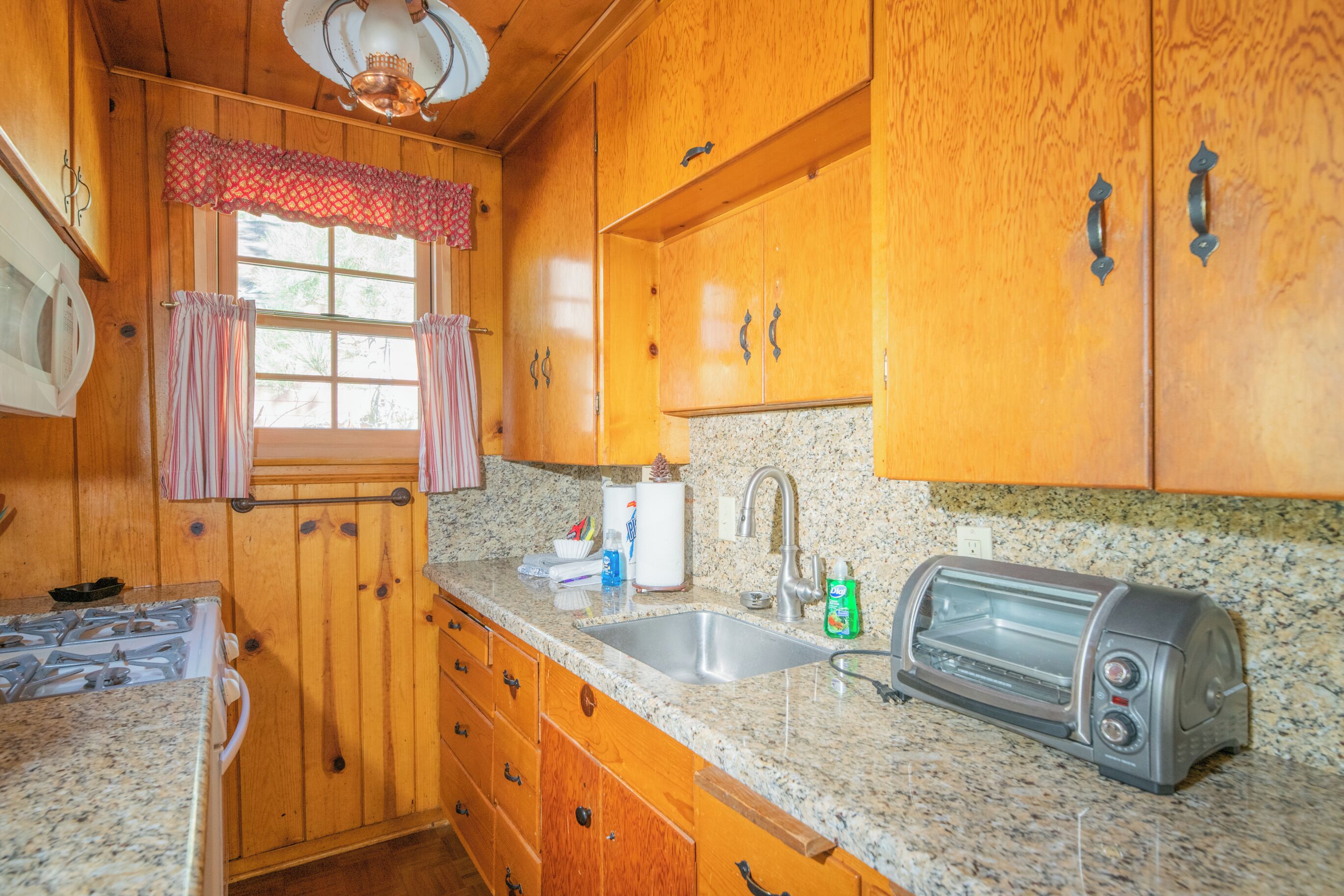 Une cuisine avec des meubles en bois, un plan de travail en granit, un évier, un grille-pain et une fenêtre avec des rideaux.