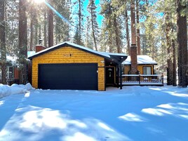 A cabin with a garage, surrounded by tall pine trees and covered in snow.