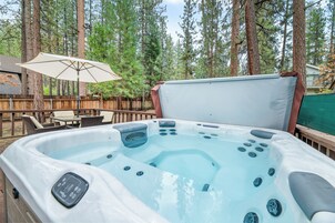 A hot tub with multiple jets, surrounded by a wooden deck and a wooden fence, with a patio umbrella and outdoor furniture in the background.