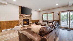 A modern living room with a stone fireplace, leather sofa, and a flat-screen TV mounted above it. - Lucky Buck by PMR, 7BR Luxury Cabin, Lake View, EV Charge, Sleeps 22 (Lake Harmony)