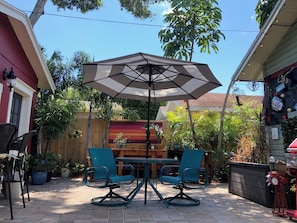 Eine Terrasse im Freien mit Tisch, zwei Stühlen, einem Sonnenschirm und verschiedenen Blumentöpfen.