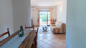 A dining area with a wooden table set for two, a sofa, and a view of a balcony with curtains.