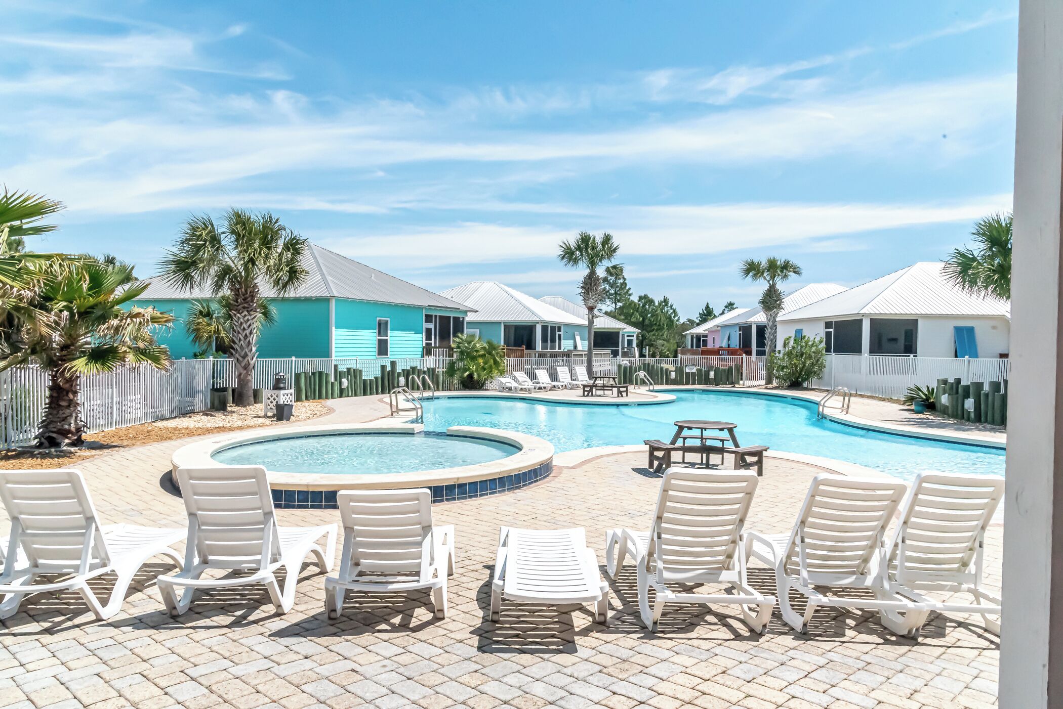 Zona de piscina con sillones de descanso y una pileta rodeada de palmeras y casas.