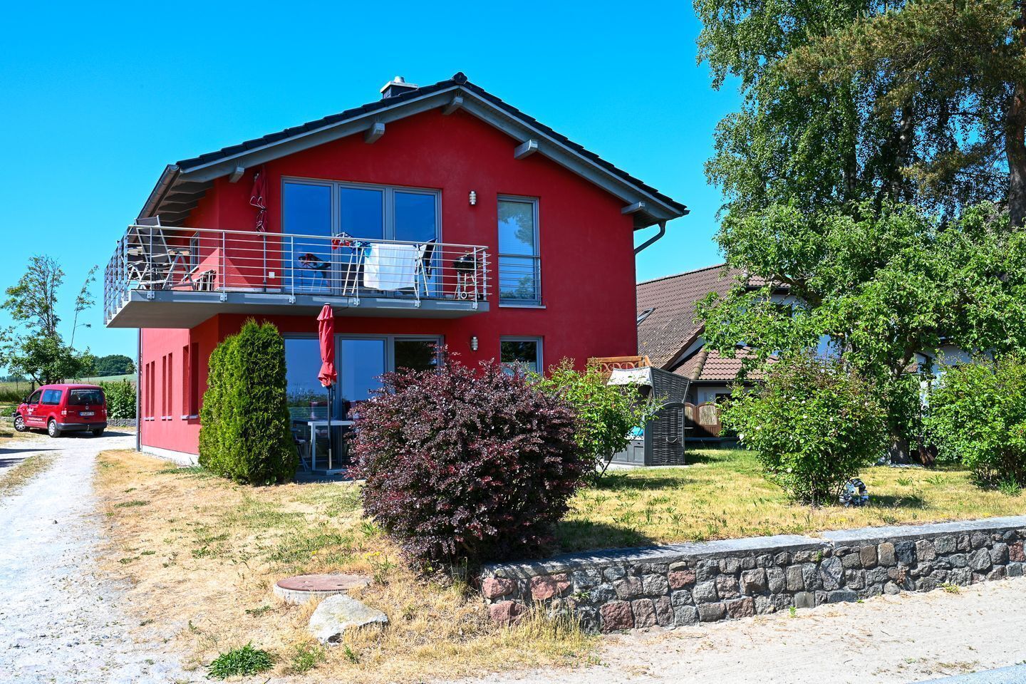 Une maison rouge à deux étages, dotée d’un balcon, d’un mur en pierre et d’un petit arbuste dans le jardin.