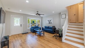 Amplia sala de estar con piso de madera, ventilador de techo, sofá azul, una mesa de centro y una escalera con escalones de madera.