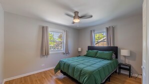 Un dormitorio con una cama, dos lámparas de noche, un ventilador de techo y dos ventanas con cortinas.