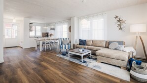 Amplia sala de estar con piso de madera, un sofá color beige, un sillón a rayas y una mesa de centro. Hay un comedor con una mesa y sillas blancas, y una cocina al fondo.