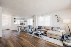 A spacious living room with a wooden floor, a beige sofa, a striped armchair, and a coffee table. There is a dining area with a white table and chairs, and a kitchen in the background.