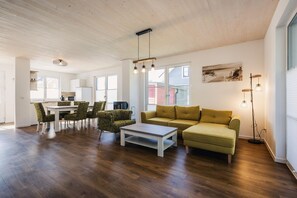 A spacious living area with a wooden floor, a yellow sofa, patterned armchairs, a dining table with chairs, and a kitchen in the background.