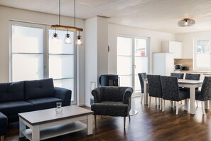 A modern living room with a sofa, armchair, dining table, and chairs. There is a kitchen area with cabinets and a microwave.