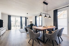 A spacious living area with a wooden dining table, grey upholstered chairs, and pendant lighting.