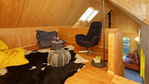 A cozy attic room with wooden walls, a black office chair, a small round table with a bottle and glass, a cowhide rug, and a yellow blanket.
