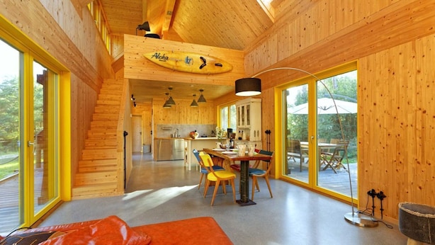 A wooden cabin interior with a dining area, kitchen, and a staircase leading to an upper level.