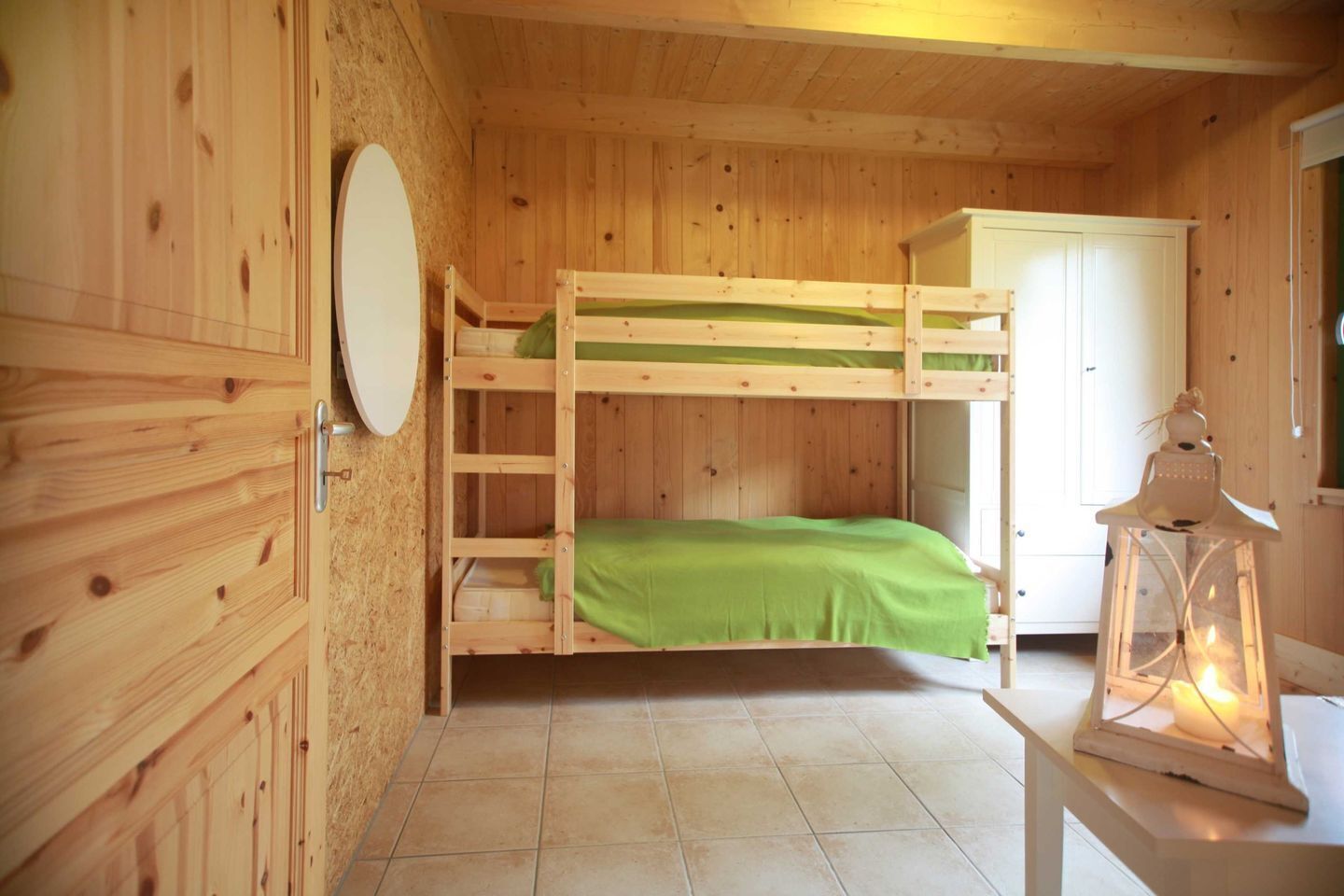 Une chambre avec un lit superposé en bois, une couette verte, une armoire blanche et une lanterne posée sur une table.