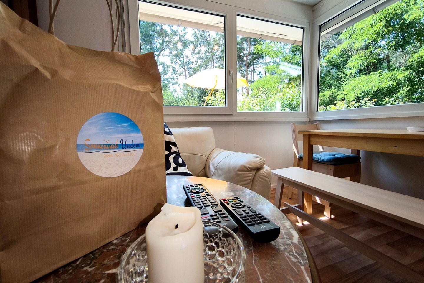 A room with a view of greenery through large windows, a sofa, a table with a candle and remote controls, and a beach-themed bag.