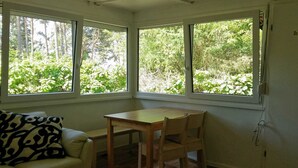 A room with a wooden table and two chairs, a sofa with a patterned pillow, and large windows with a view of greenery. - Gemütliches, 30 qm Großes Ferienhaus für 4 Personen. - Minna-busch-weg (Loddin)