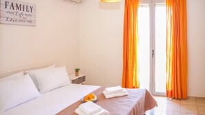 A bedroom with a bed, pillows, a bedside table with a plant, a sign on the wall, and orange curtains. - Family Villa Hera With Sea View (Mylopotamos)