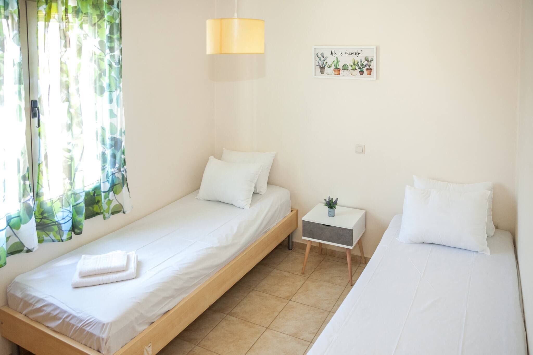 A small, clean room with two single beds, a bedside table with a lamp, and a wall-mounted plant.