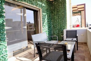 A balcony with a glass door, a green leafy wall, a table with chairs, and a TV.