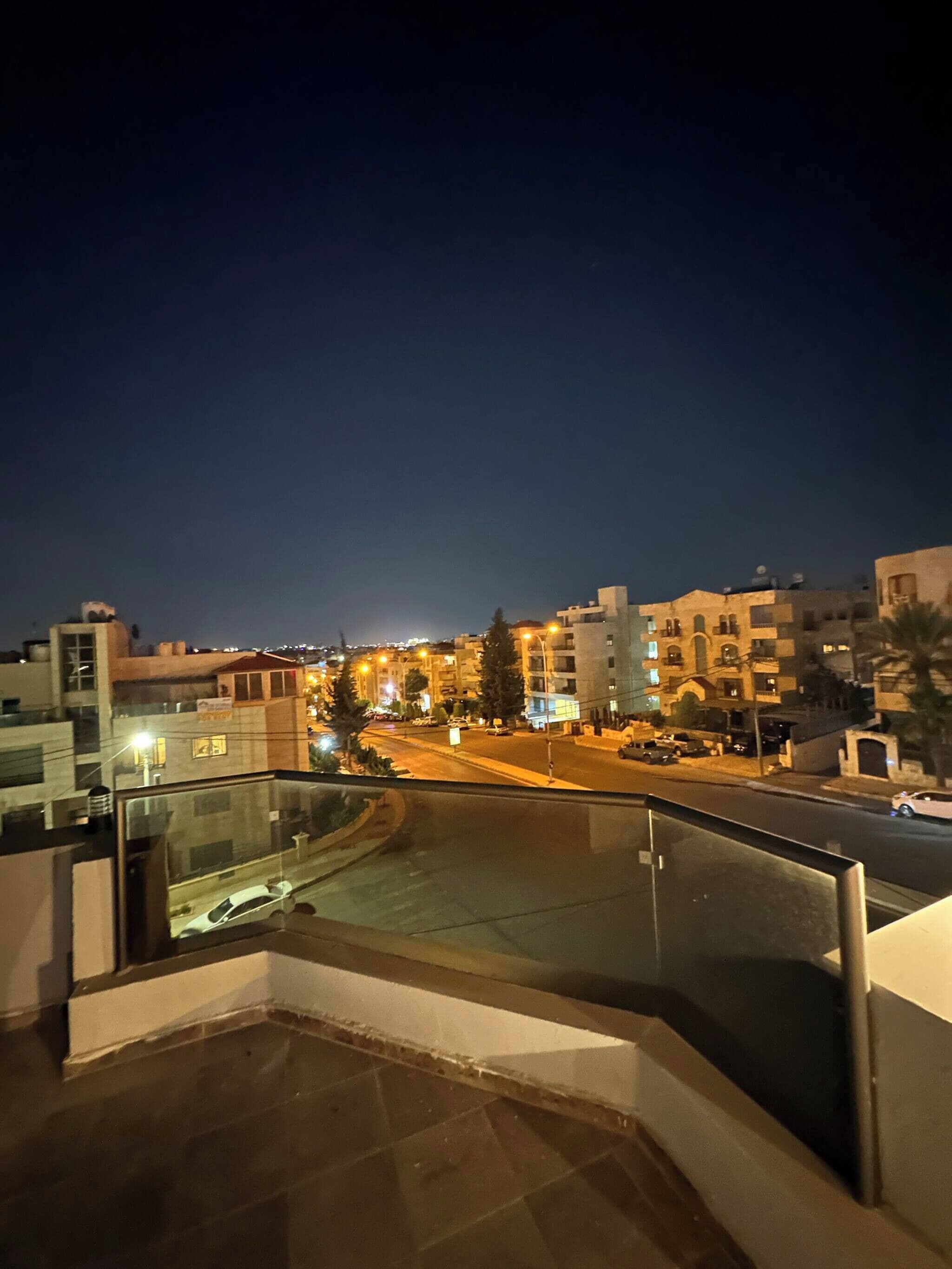 A nighttime cityscape with buildings, a street, and a pedestrian walkway.