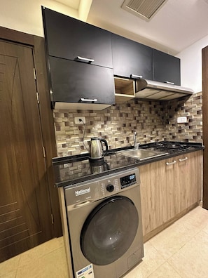 Compact kitchen with stainless steel washing machine, black countertop, and brick-patterned backsplash.