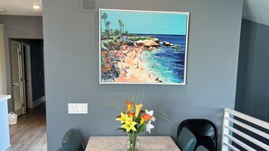 A dining area with a table, chairs, a vase of flowers, and a beach painting on the wall.