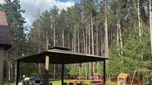 A covered patio with outdoor furniture, a small playhouse, and a swing set in a forested area.