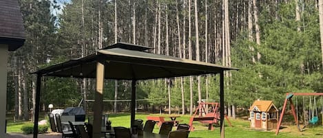 A covered patio with outdoor furniture, a small playhouse, and a swing set in a forested area.