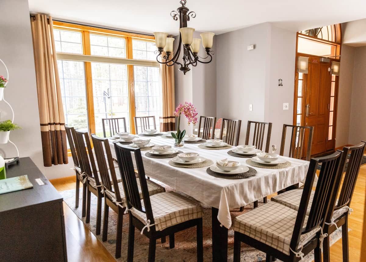 A dining room with a table set for eight, surrounded by chairs with checkered cushions, a chandelier, and a window with curtains.