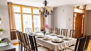 A dining room with a table set for eight, surrounded by chairs with checkered cushions, a chandelier, and a window with curtains.
