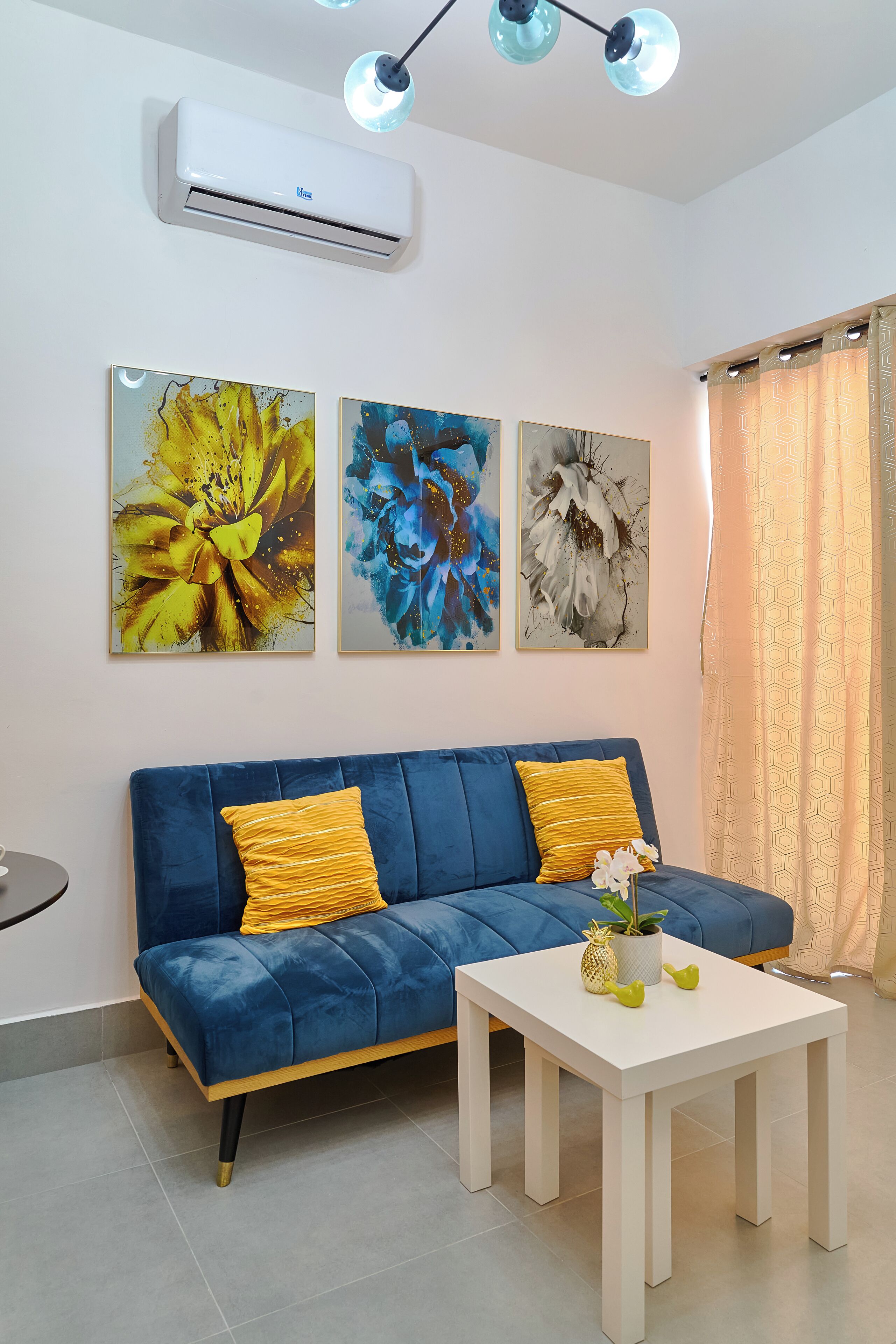 A living room with a blue sofa, two yellow cushions, a white coffee table, and three framed floral artworks on the wall.