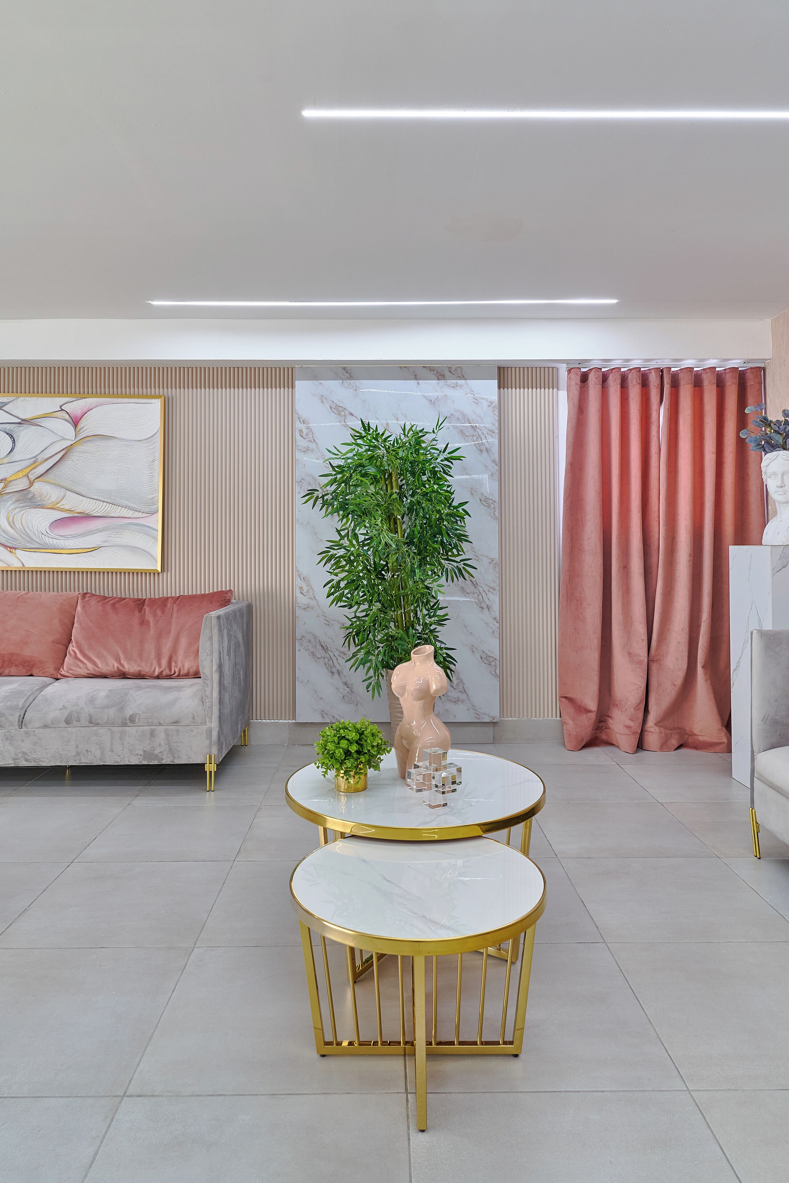 Una sala de estar moderna con un sofá gris, una mesa redonda con patas doradas, una planta en maceta y una pared de mármol.