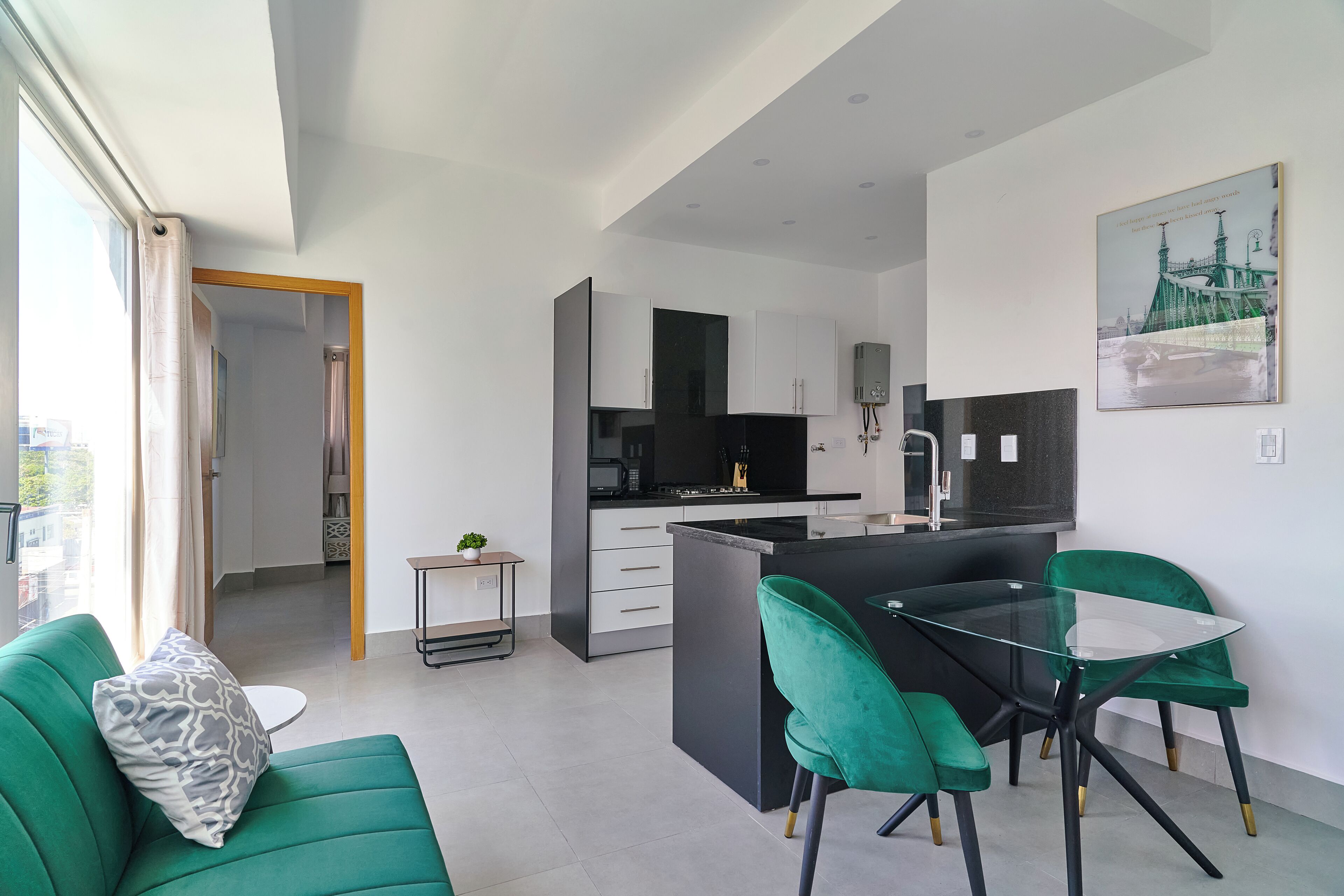 Una cucina moderna con mobili bianchi, un piano di lavoro nero e un'isola in vetro. Nell'area soggiorno, un divano verde con un cuscino decorato.