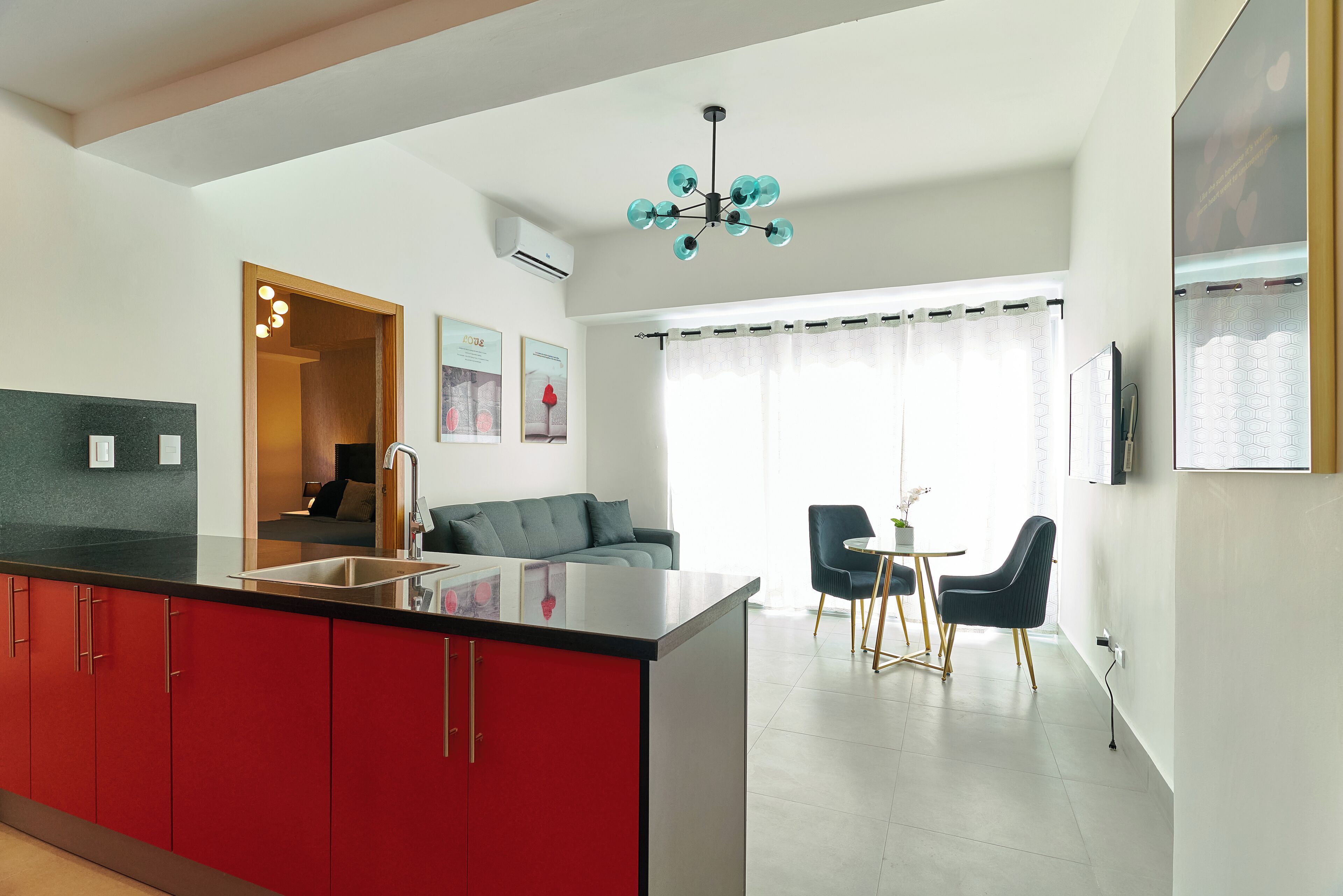 A modern kitchen with red cabinets, a black countertop, and a built-in sink. There is a dining area with a table and chairs, and a sofa in the background.