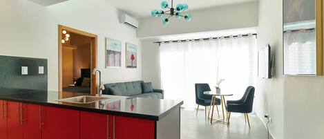 A modern kitchen with red cabinets, a black countertop, and a built-in sink. There is a dining area with a table and chairs, and a sofa in the background.