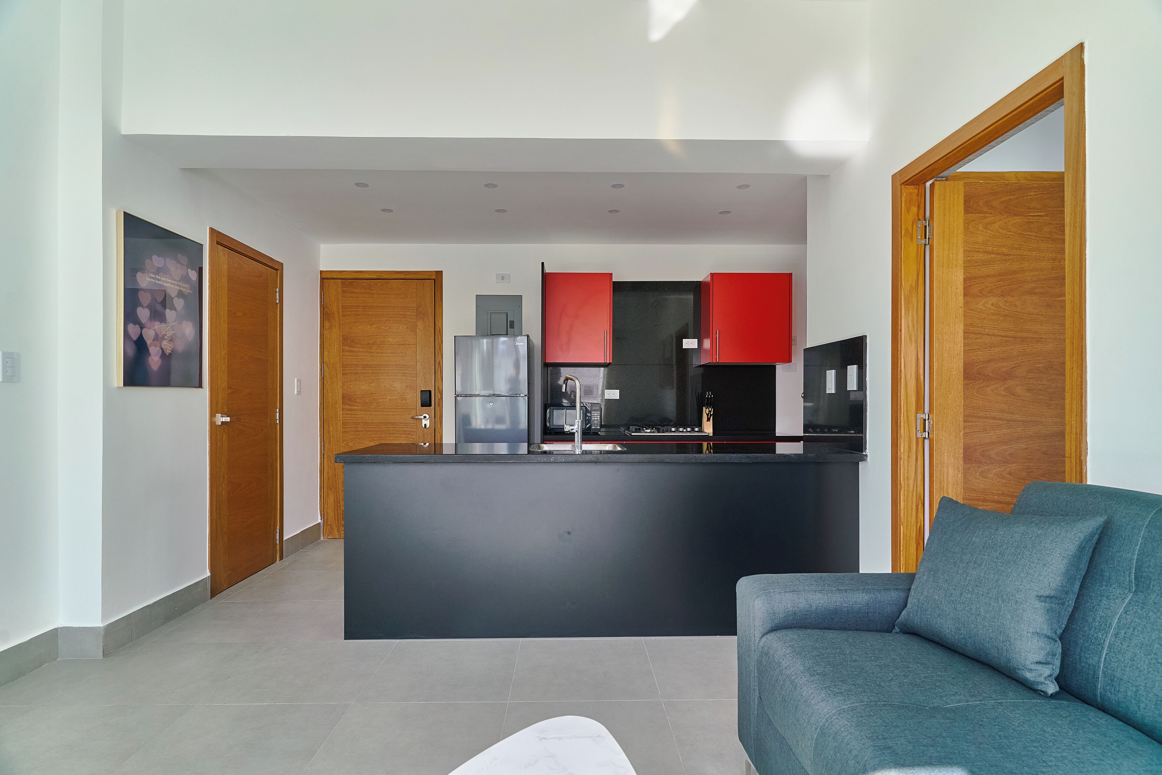 A modern kitchen with a central island, red cabinets, and a black countertop. There is a grey sofa in the living area.