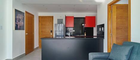 A modern kitchen with a central island, red cabinets, and a black countertop. There is a grey sofa in the living area.