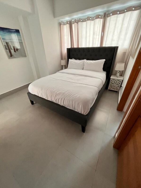 Una cama bien hecha con un cabecero oscuro en una habitación con paredes de color claro y suelo de baldosas.