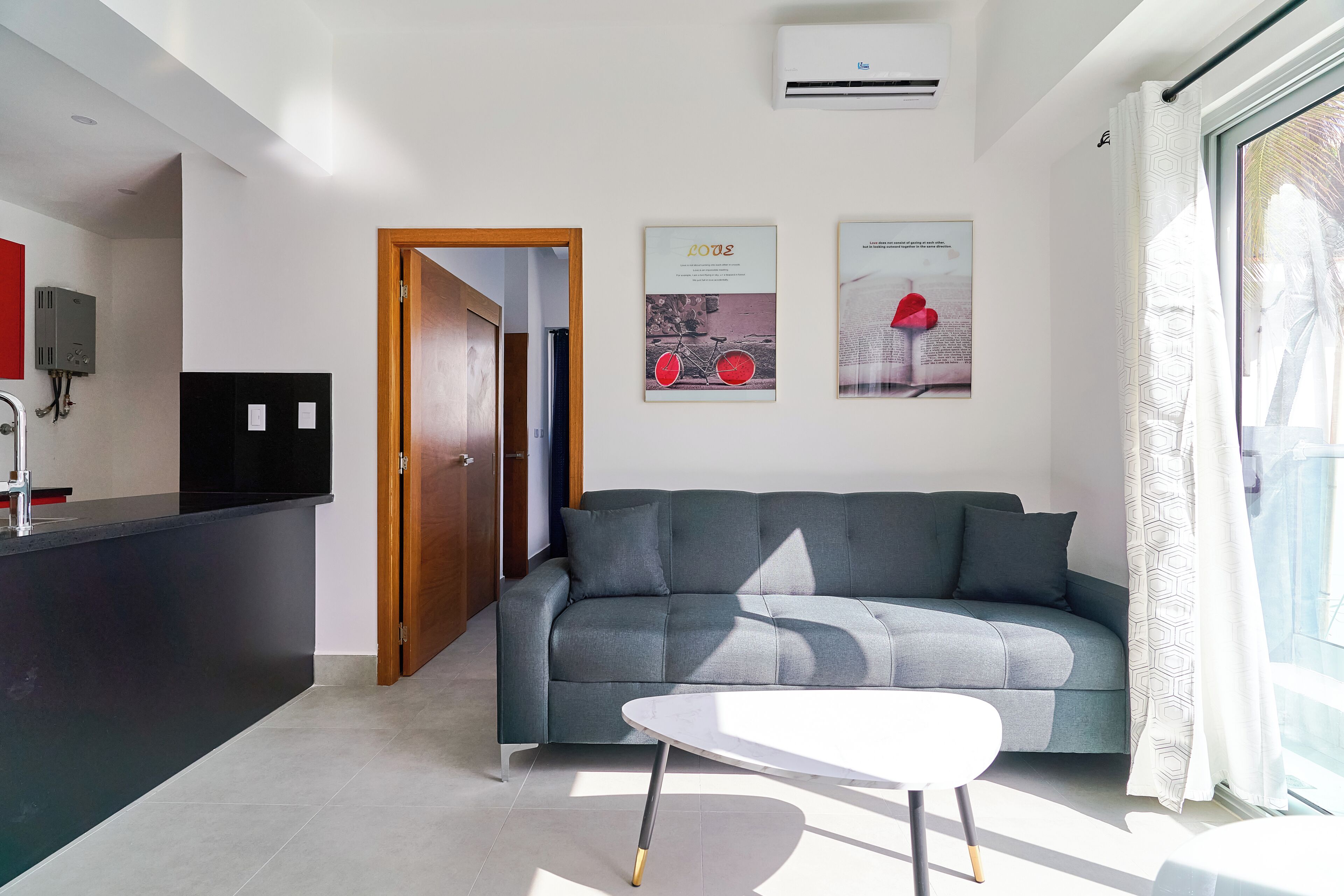 Un salon moderne avec un canapé gris, une table basse blanche et un coin cuisine doté d’un plan de travail noir et d’un appareil électroménager rouge.