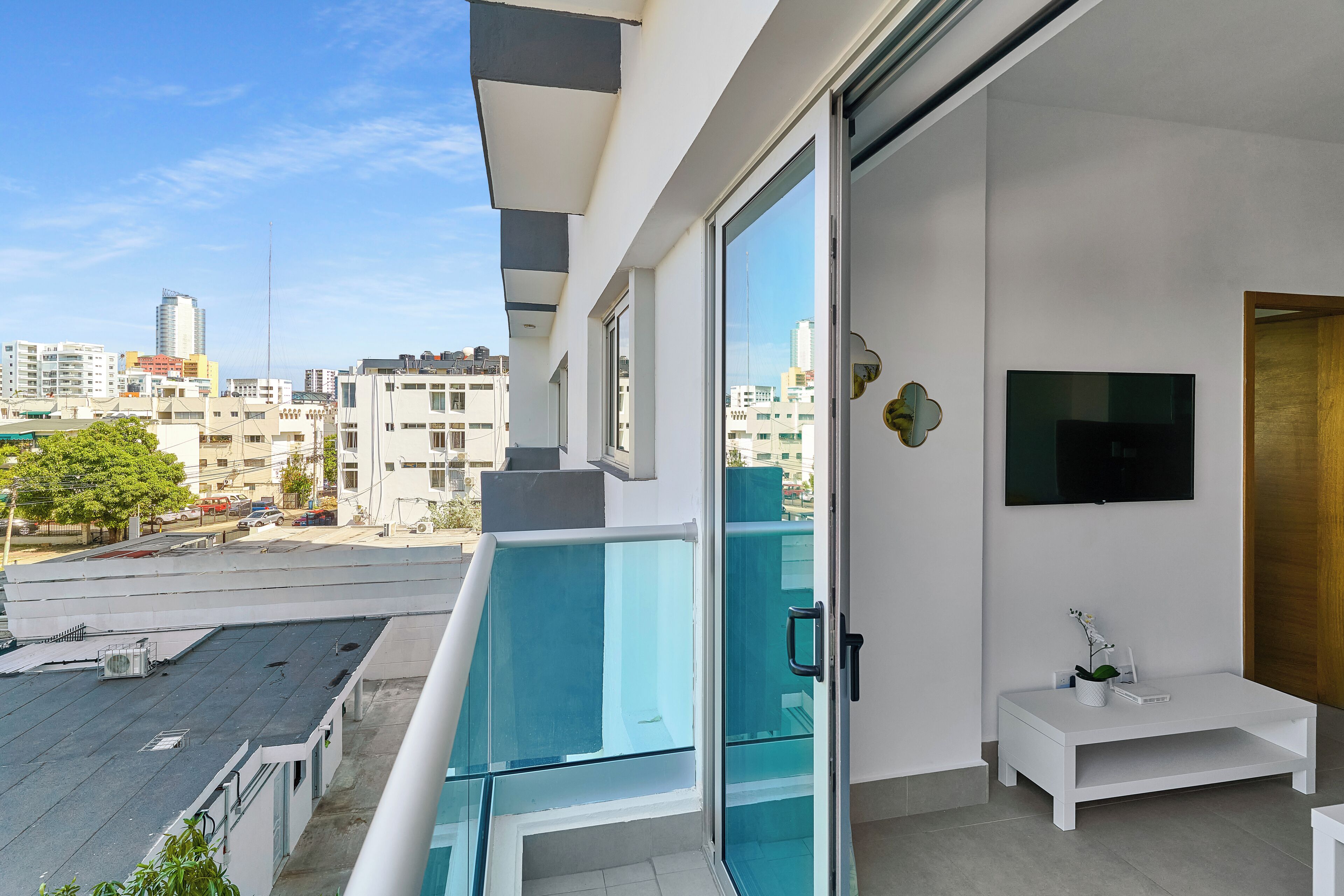 A modern apartment with a balcony overlooking a cityscape, a flat-screen TV mounted on the wall, and a small white side table with a plant.